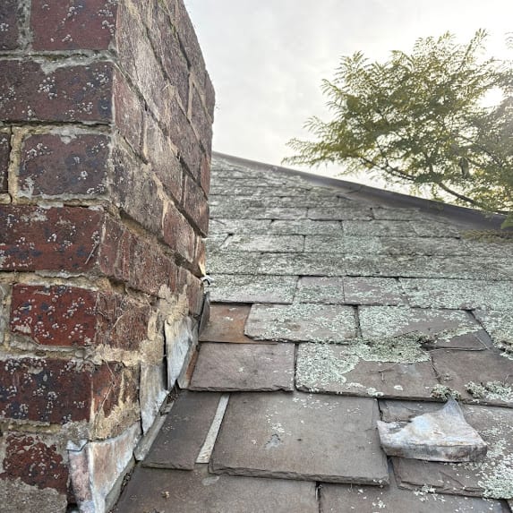 Lichen-covered slate against a brick chimney, with stepped lead flashing at the junction